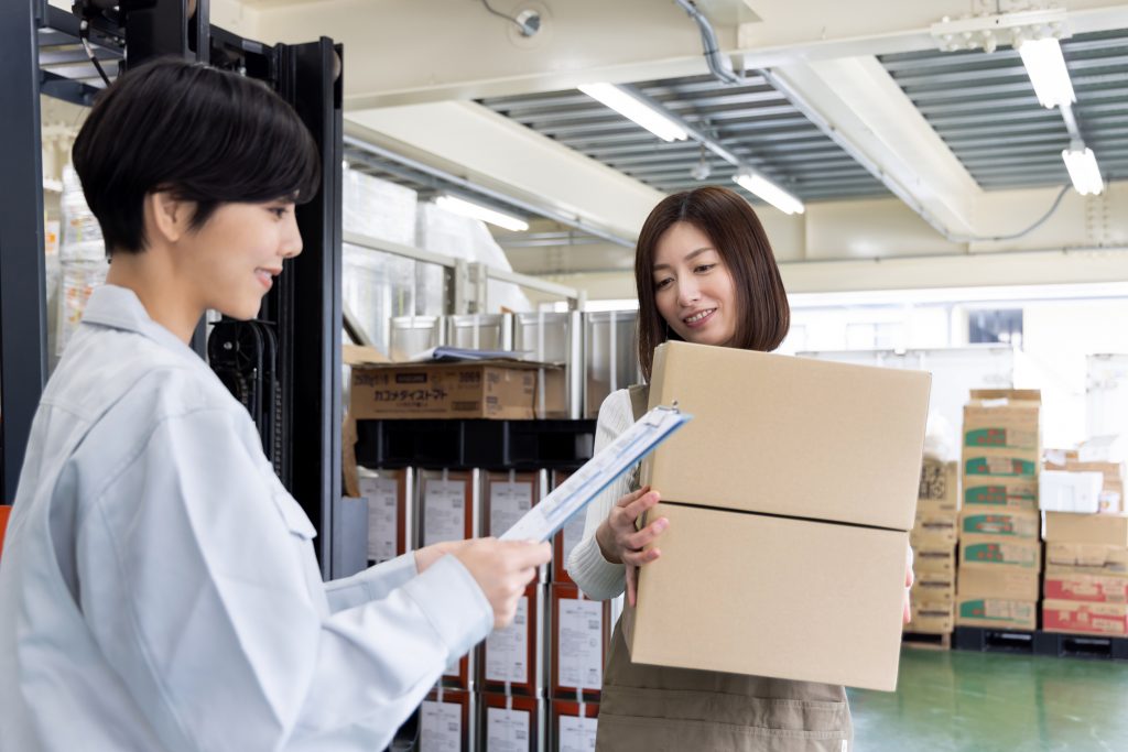 倉庫で段ボールを持つビジネスパーソンと資料を持ちながら話すビジネスパーソン