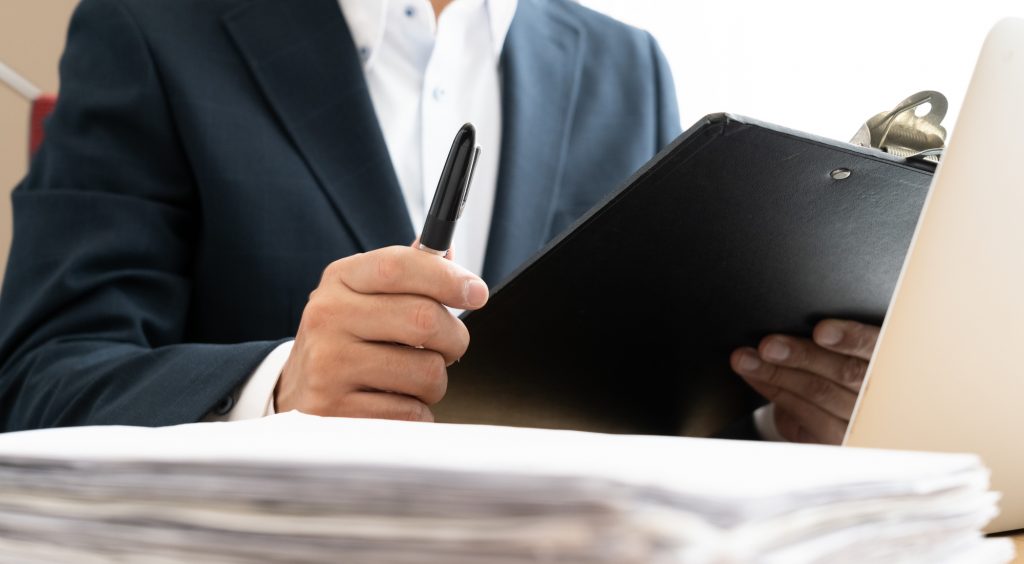 パソコンと資料が置かれたデスクに座り、ペンを片手に資料を見るビジネスパーソン