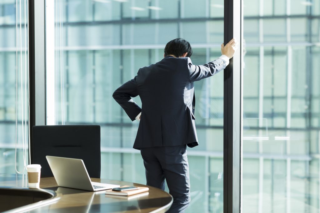 窓に手をつきうつむき悩むビジネスパーソン