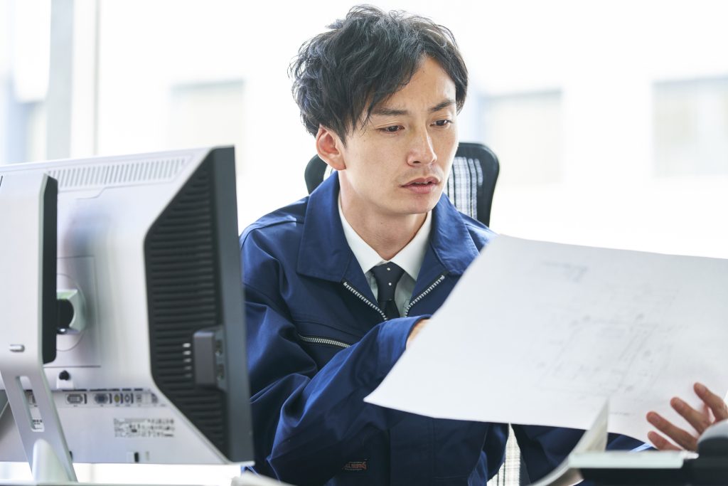 パソコンが置いてあるデスクに座り、資料を見ながら考える若いビジネスパーソン