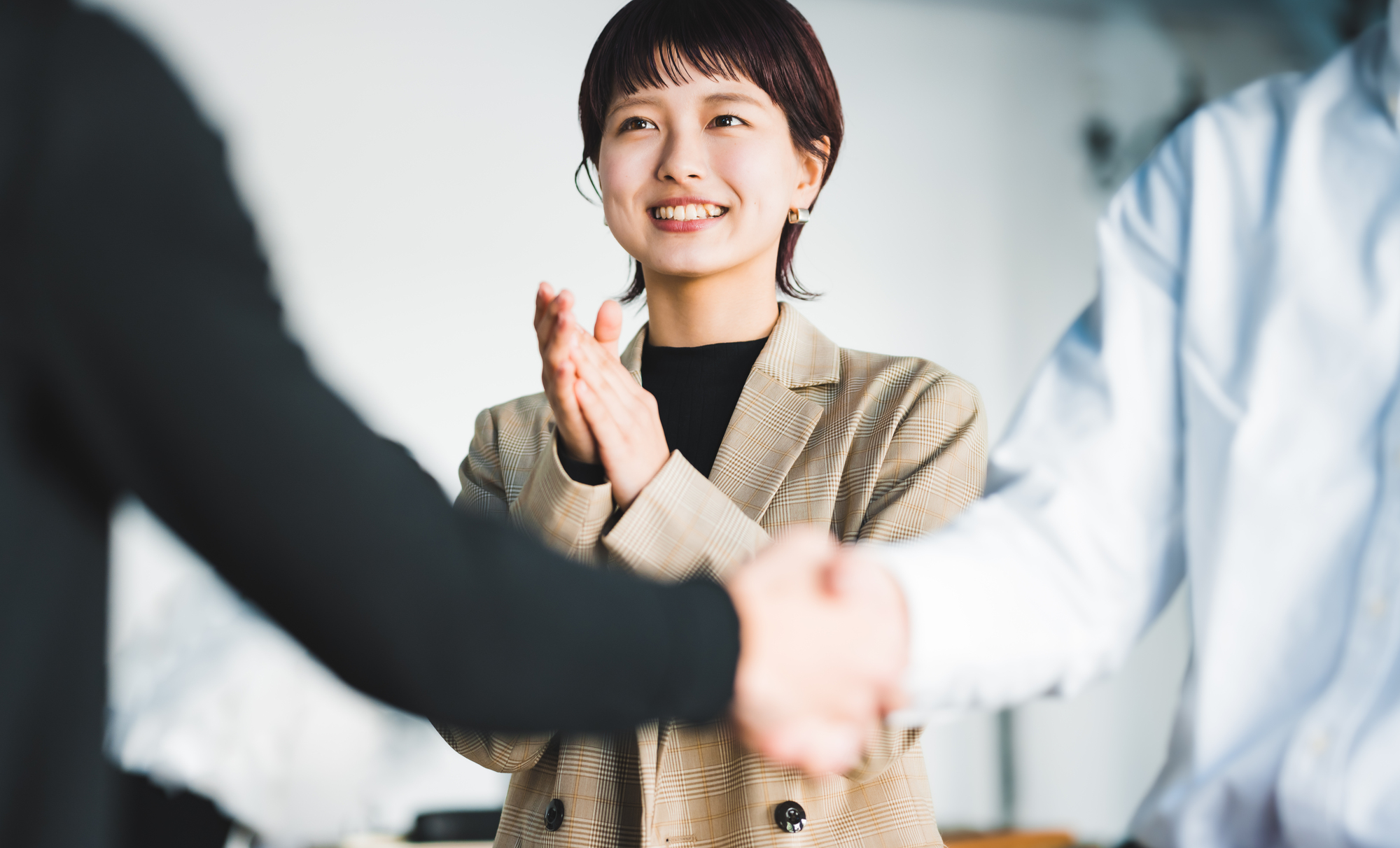 握手する2人のビジネスパーソンを前に笑顔で拍手をする女性