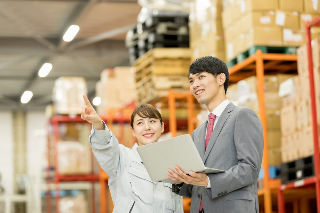 倉庫で指を指しながらほほ笑むビジネスパーソンとパソコンを片手にほほ笑むビジネスパーソン