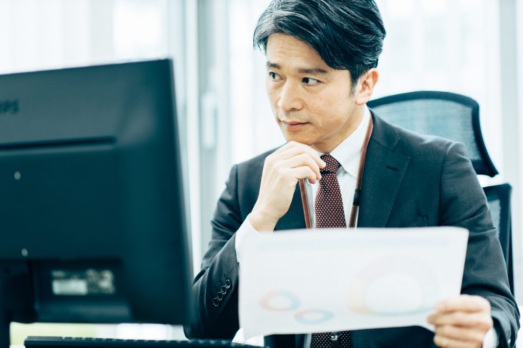 パソコンを前に資料を手にして考えるビジネスパーソン