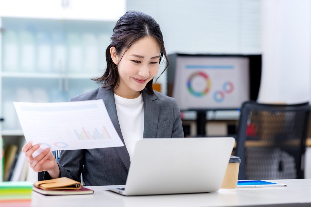 デスクで資料を片手にパソコン作業をしながらほほ笑むビジネスパーソン