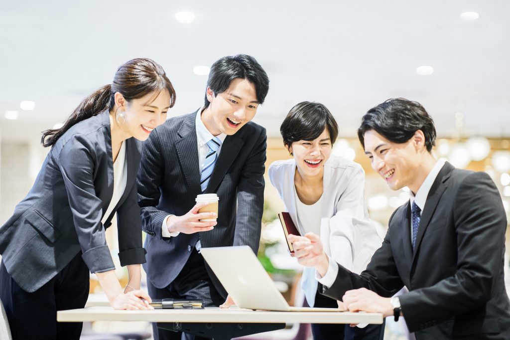 デスクでパソコンを見る男性を3人のビジネスパーソンが笑顔で囲む