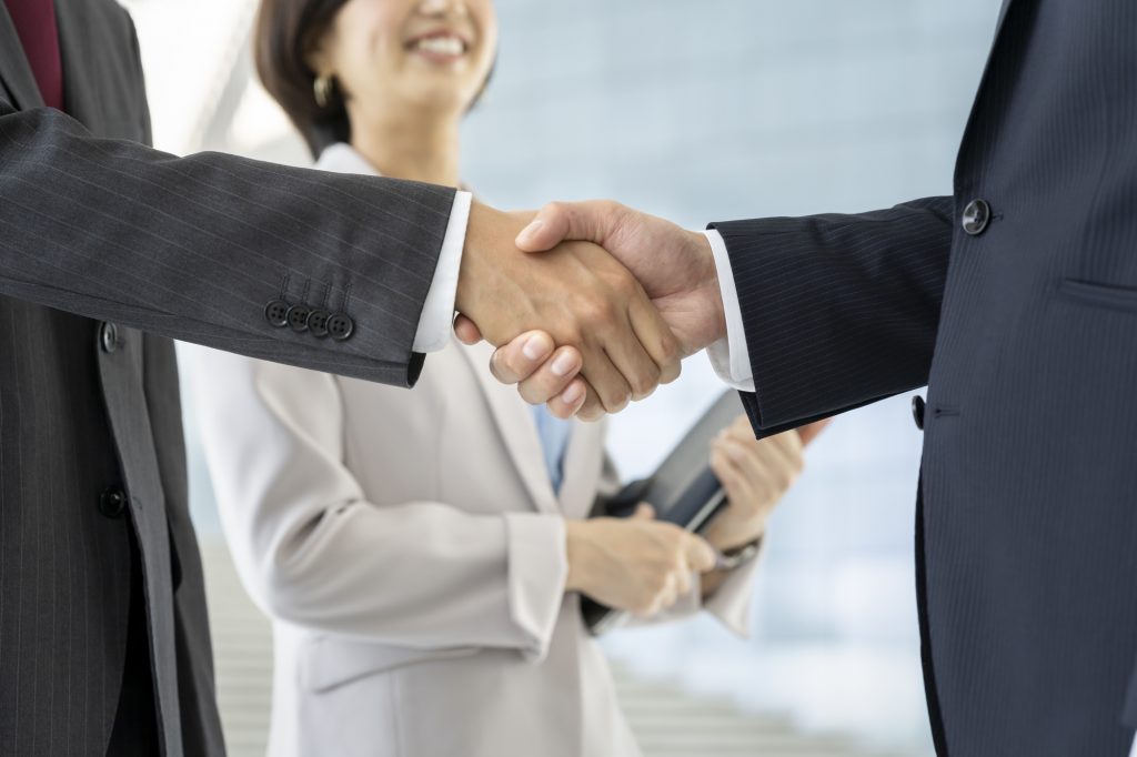 握手をする2人のビジネスパーソンの手元と、それを見ながらほほ笑む1人のビジネスパーソン
