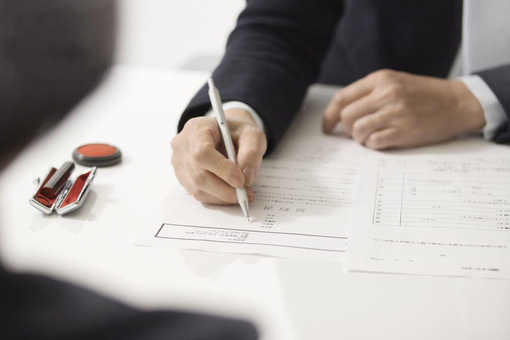 印鑑が置かれたデスクで書類に記入する男性の手元