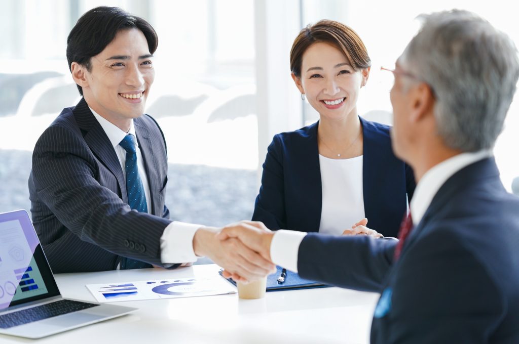 握手する2人のビジネスパーソンとそれを見てほほ笑むビジネスパーソン