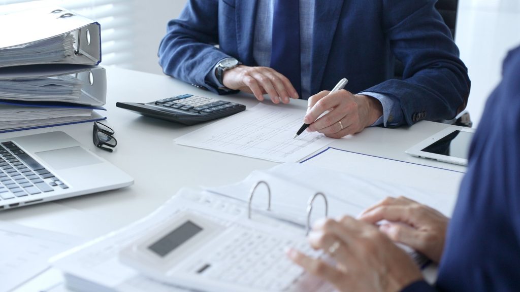 資料やパソコン、電卓の置かれたデスクで会議するビジネスパーソン