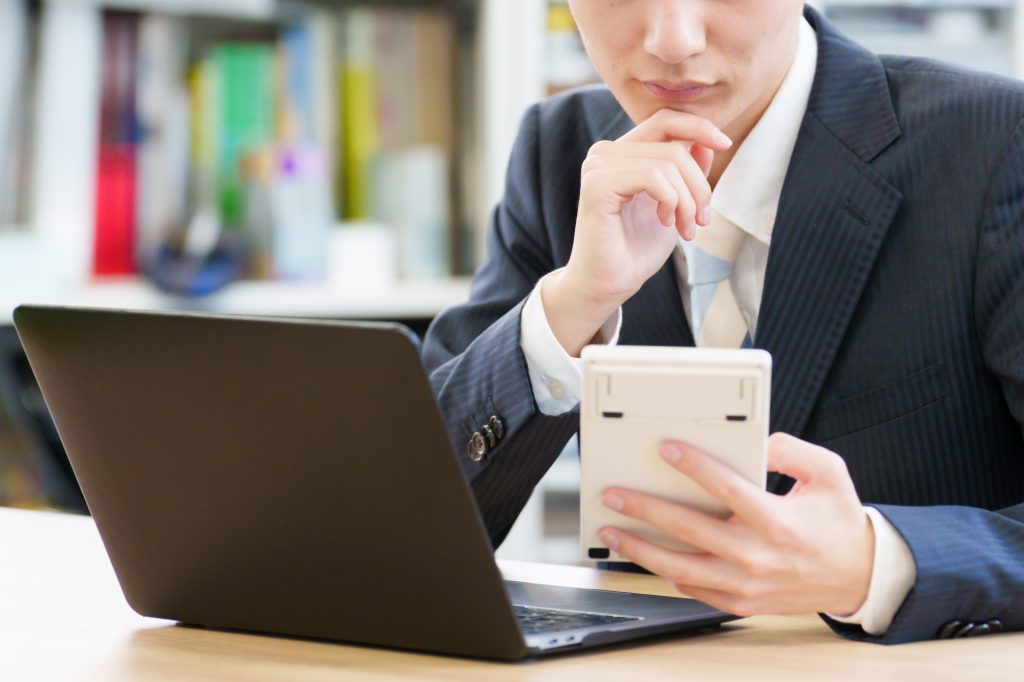 パソコンが置かれたデスクで電卓を見ながら悩むビジネスパーソン