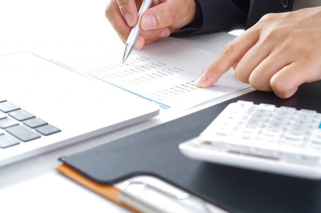 パソコンと電卓が置かれたデスクで、資料を見ながら作業するビジネスパーソンの手元