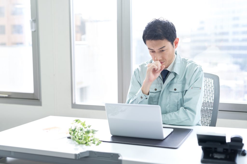 デスクでパソコンを見ながら考える作業着を着たビジネスパーソン