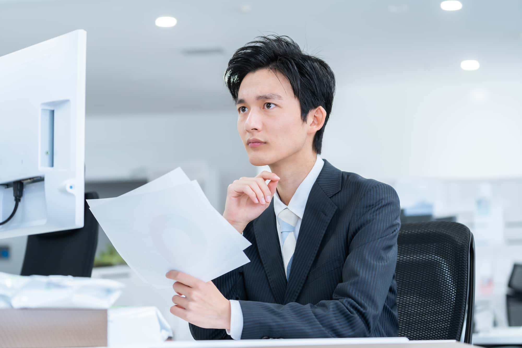 オフィスで資料を手にしながらパソコンを見るビジネスパーソン