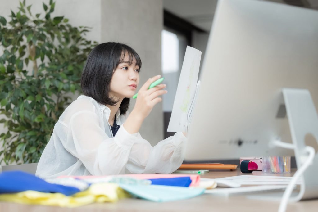 パソコンが置かれたデスクで資料を見ながら考えごとをするビジネスパーソン
