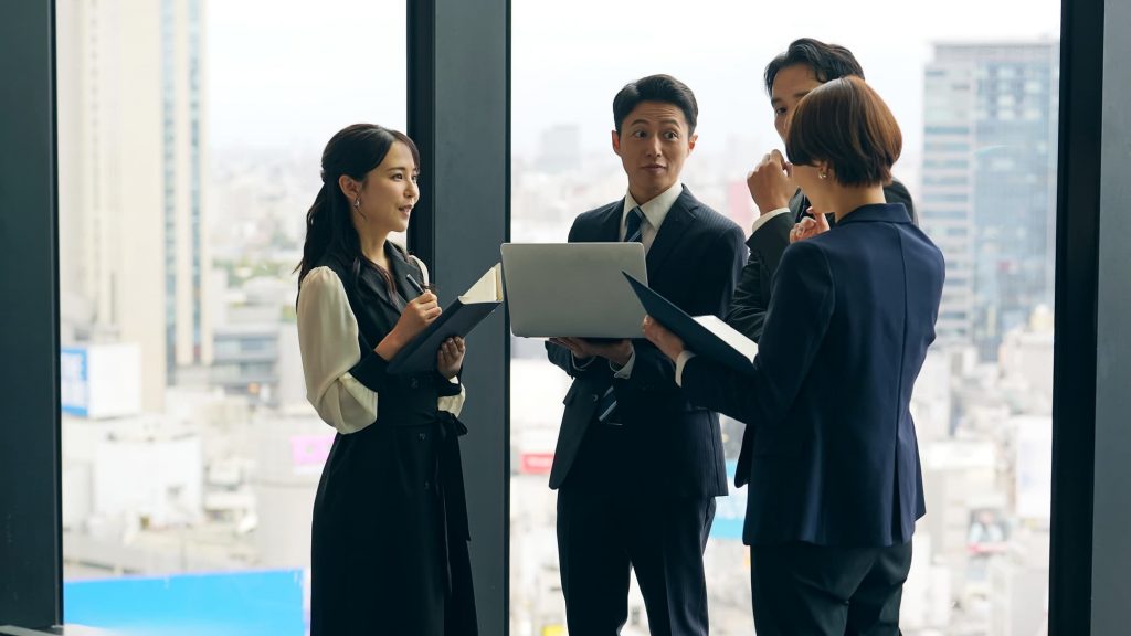 オフィスで資料とパソコンを見ながら話す4人のビジネスパーソン
