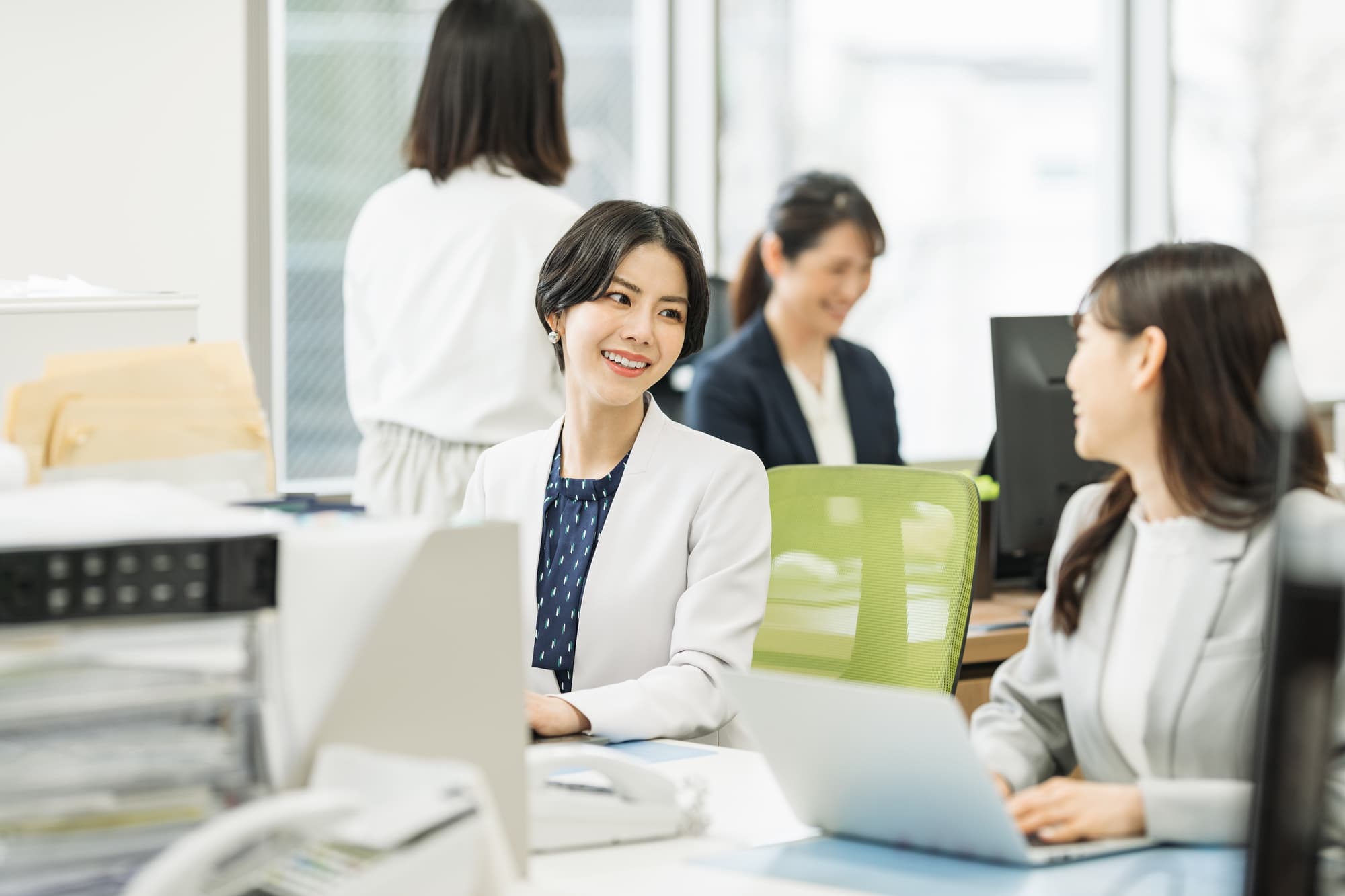 パソコン作業をしながらデスクで会話する2人のビジネスパーソン